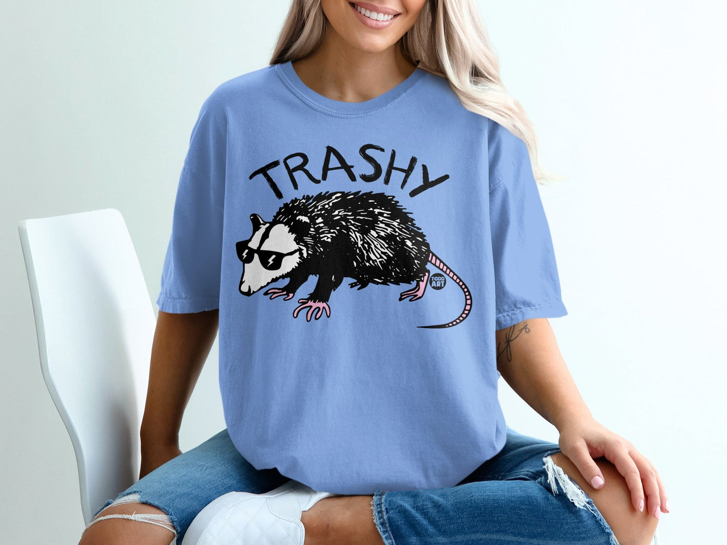 Opossum wearing sunglasses with "TRASHY" text on a blue t-shirt.