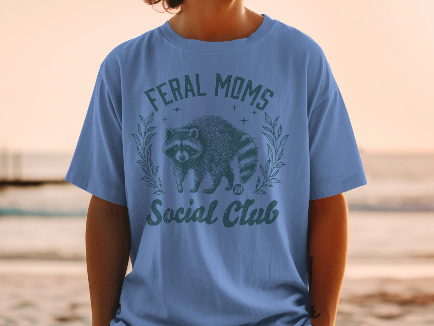 Blue t-shirt with "Feral Moms Social Club" and raccoon graphic.