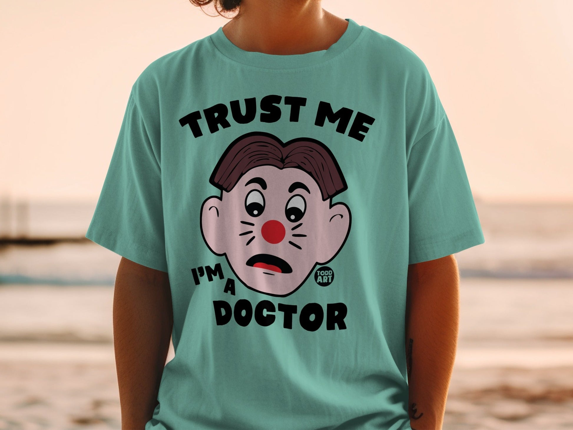 Teal t-shirt with cartoon clown face and text "TRUST ME I'M A DOCTOR".