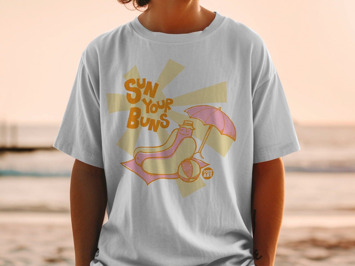 T-shirt featuring a hot dog with a pink umbrella on a beach towel.