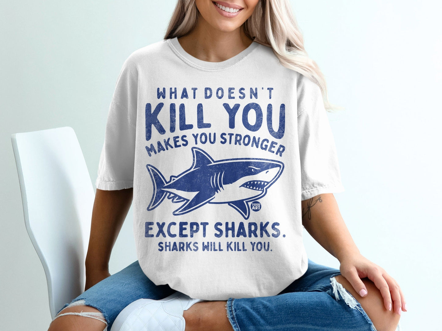White t-shirt with blue text and shark graphic. "What doesn't kill you makes you stronger except sharks."