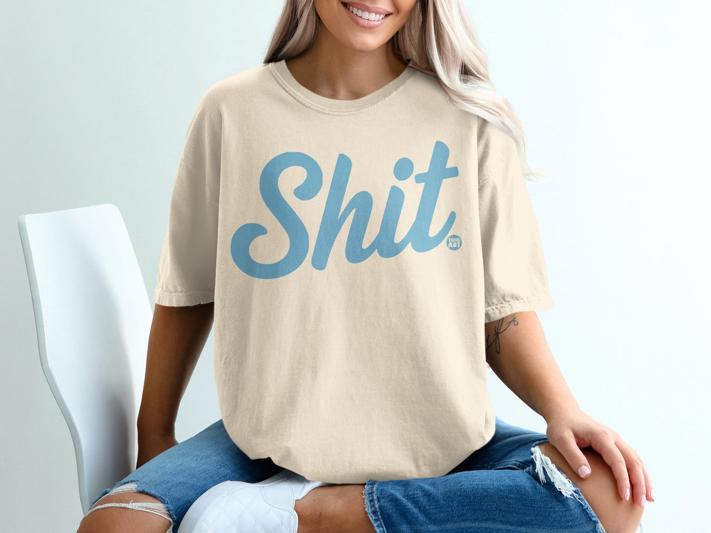 Cream t-shirt with light blue "Shit Happens" graphic, worn by a person sitting.
