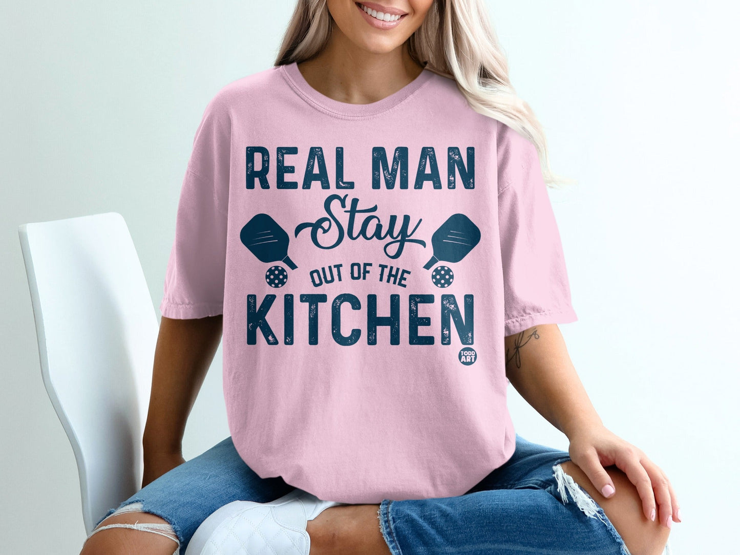 Pink t-shirt with "Real Man Stay Out Of The Kitchen" text and pickleball paddles.