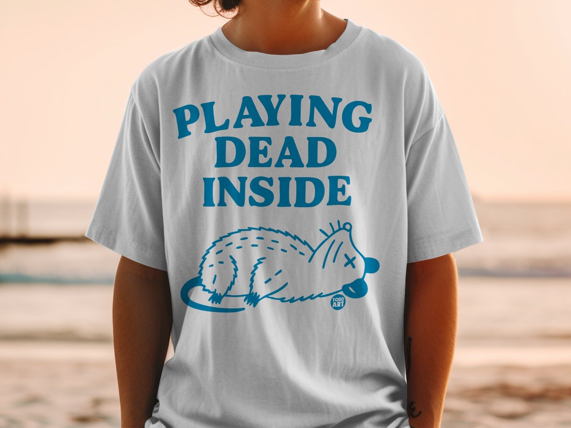 White t-shirt with "PLAYING DEAD INSIDE" text and a blue opossum illustration.