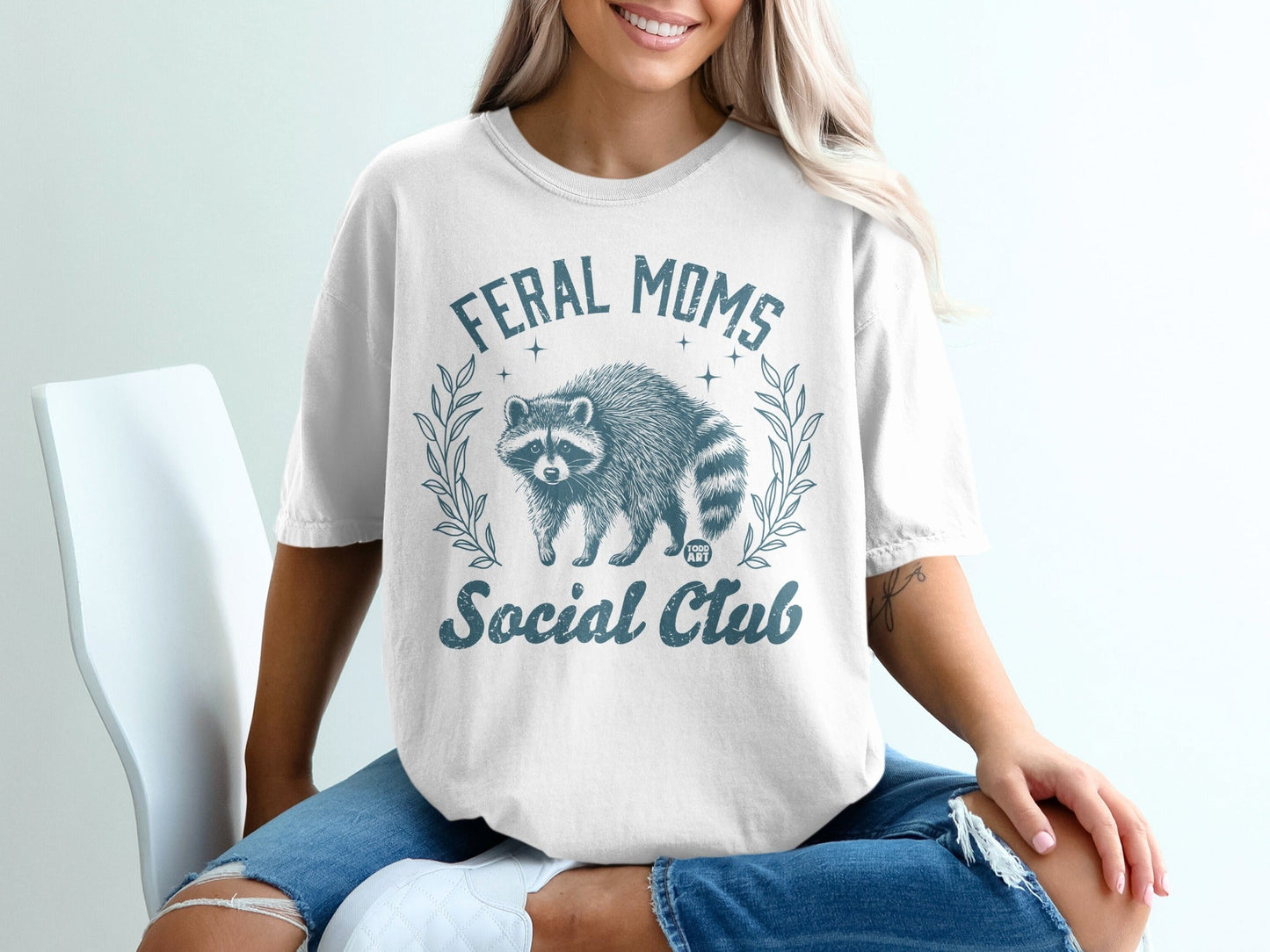 White t-shirt with raccoon and "Feral Moms Social Club" text.