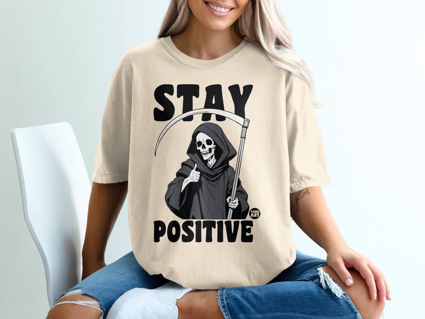 Grim reaper giving thumbs up with "STAY POSITIVE" text on a light t-shirt.