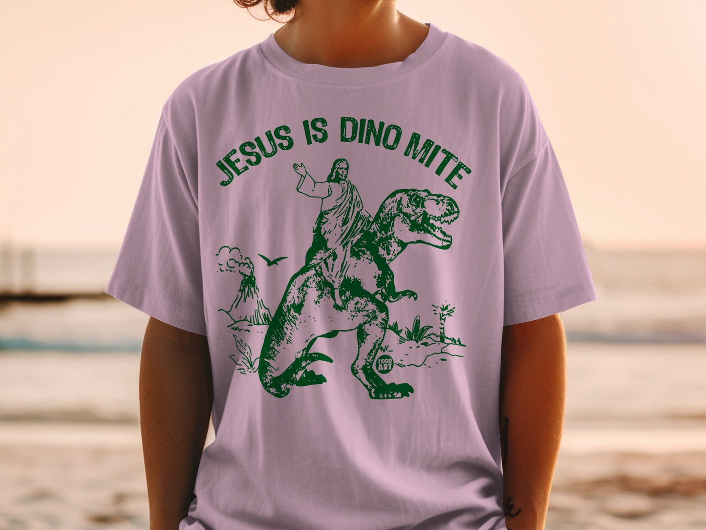 Person wearing a light purple t-shirt with a green graphic of Jesus riding a dinosaur.