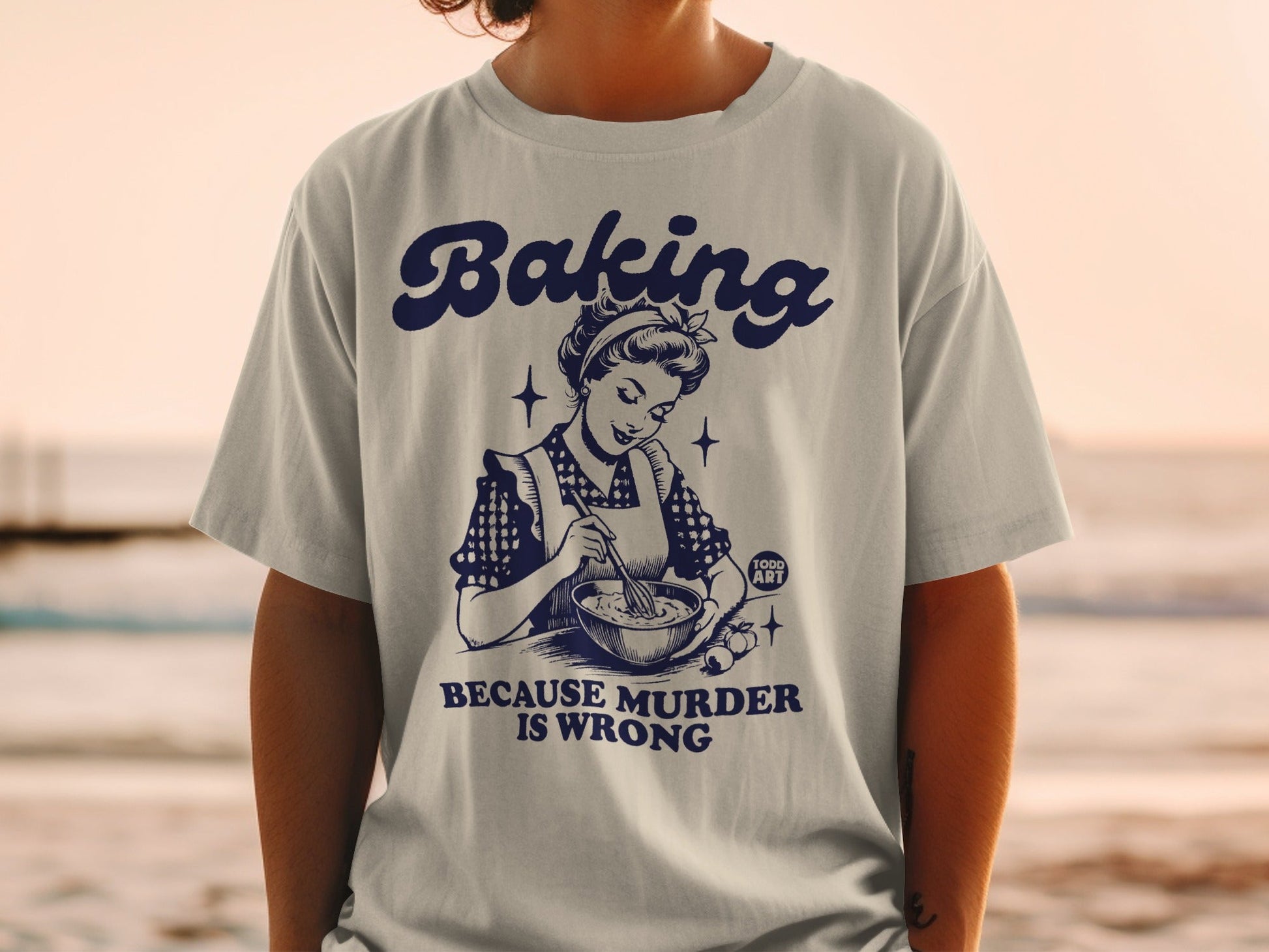 White t-shirt with navy graphic of woman baking and text "Baking Because Murder Is Wrong".