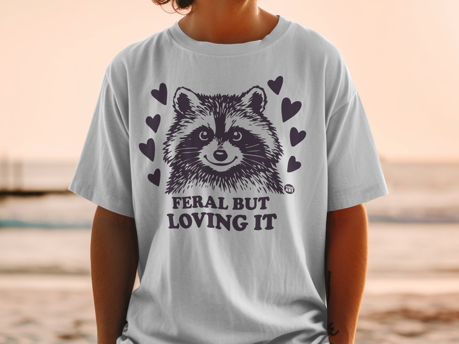 White t-shirt with raccoon graphic and text "Feral But Loving It".