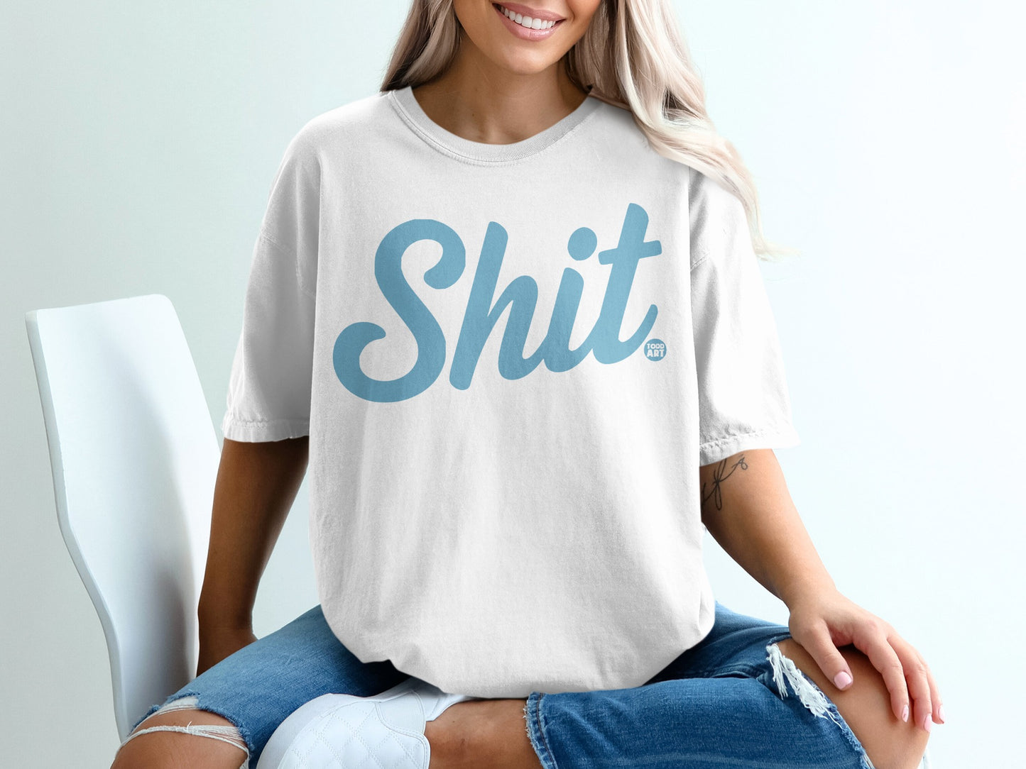 White t-shirt with large light blue "Shit" graphic, worn by a seated person.