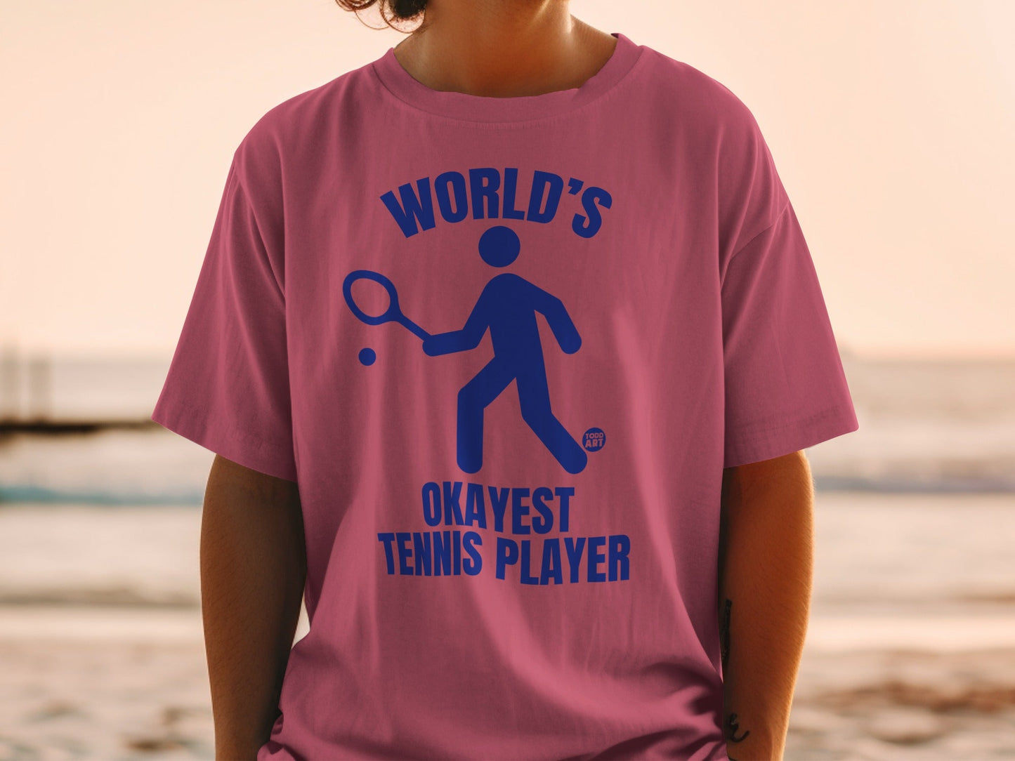 Pink t-shirt with blue graphic of a stick figure playing tennis and text "WORLD'S OKAYEST TENNIS PLAYER".