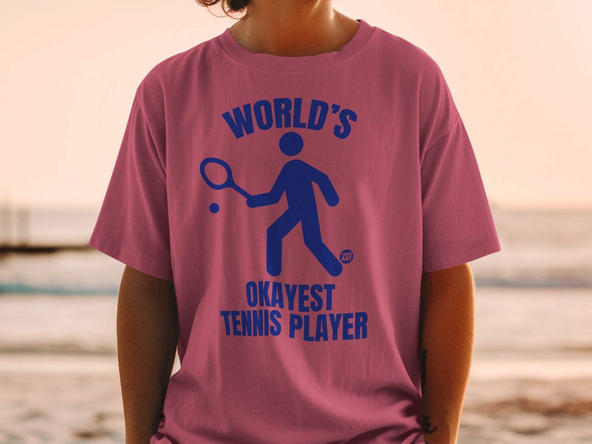 Pink t-shirt with blue graphic of a stick figure playing tennis and text "WORLD'S OKAYEST TENNIS PLAYER".